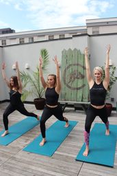 Three women doing rooftop yoga on blue mats in warrior lunge on an urban wooden deck terrace with potted palms and a weathered wooden backdrop — outdoor rooftop yoga class.
