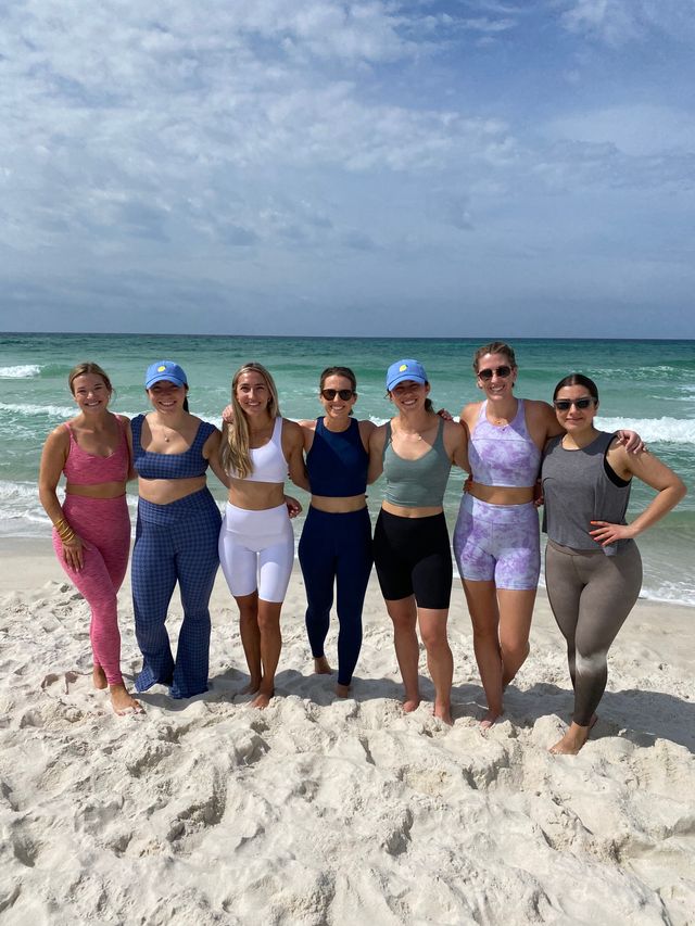 Seven friends in colorful activewear pose arm-in-arm on a white sand beach with turquoise ocean and a partly cloudy sky.