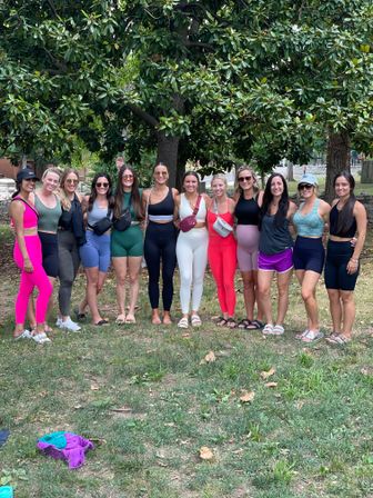 Outdoor fitness group photo of 12 women in colorful leggings and athletic tops standing arm-in-arm under a large shade tree in a neighborhood park