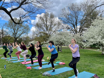 Group of women practicing outdoor yoga on mats in a city park, balancing in chair pose on a green lawn beneath white spring blossoms and a partly cloudy blue sky.