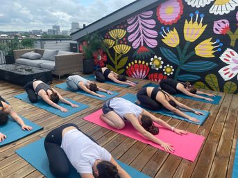 Rooftop yoga class on a wooden deck with students in child’s pose in front of a vibrant floral mural and distant city skyline, bright urban outdoor workout scene