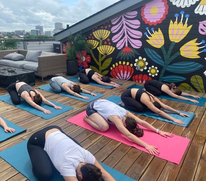 Rooftop yoga class on a wooden deck with students in child’s pose in front of a vibrant floral mural and distant city skyline, bright urban outdoor workout scene