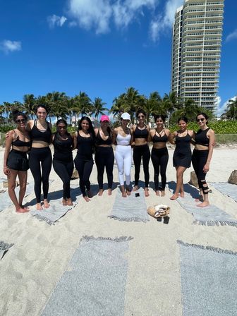 Group of women in activewear posing on yoga mats on a sunny South Florida beach with palm trees and a tall oceanfront condo under a bright blue sky.