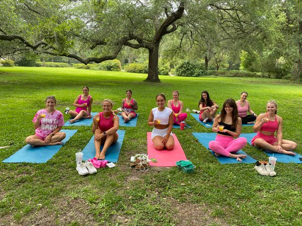 Group of women in bright pink athletic wear sitting on yoga mats under a large oak tree on a green park lawn, smiling and raising juice glasses after an outdoor yoga session.