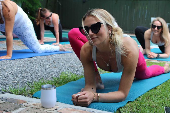 Outdoor backyard yoga session with women on blue mats — woman in heart-shaped sunglasses and pink leggings holds a low lunge while others smile, a white tumbler sits on the patio edge.