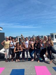 Rooftop yoga class of women in leggings and tanks striking tree and prayer poses on colorful mats under a bright blue sky, celebrating with one person holding a champagne bottle against an urban skyline.