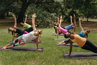 Outdoor group yoga class in a park practicing side-plank poses on mats — diverse women in colorful activewear and sunglasses under large shade trees, group fitness scene