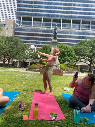 Woman popping champagne on a pink yoga mat in a downtown urban park, foam spraying as friends capture the celebratory moment by a statue with office buildings in the background.