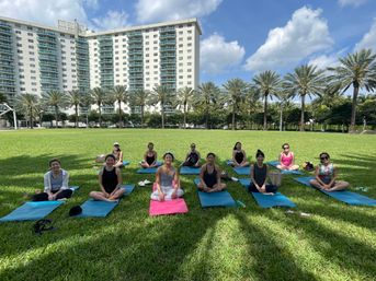 Group practicing outdoor yoga on colorful mats on a sunny green lawn, framed by palm trees and tall waterfront condos under a blue sky
