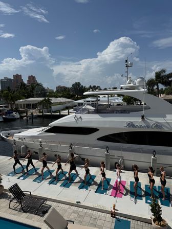 Group outdoor yoga class on a waterfront marina dock beside a white luxury yacht, palm trees and high-rise condos under a sunny blue sky.