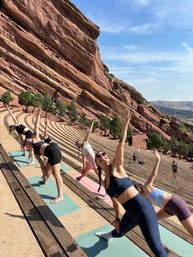 Group outdoor yoga on mats at a cliffside red sandstone amphitheater in Colorado — participants stretch and reach skyward under a sunny blue sky.