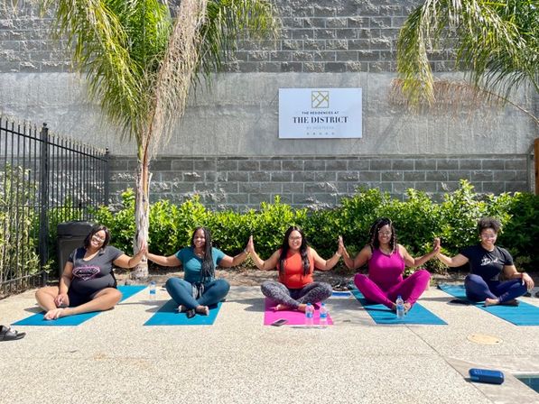 Five women seated on colorful yoga mats in a sunny palm-lined courtyard, holding hands and smiling during an outdoor yoga session.