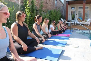 Sunlit poolside yoga class on a residential patio — women seated on mats wearing heart-shaped sunglasses.