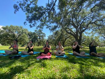 Six people in an outdoor yoga class seated cross-legged on colorful mats under large oak trees in a sunny park, hands together in a prayer pose for meditation.