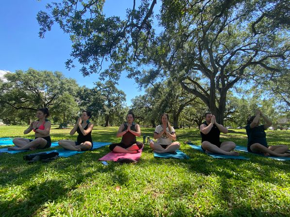 Six people in an outdoor yoga class seated cross-legged on colorful mats under large oak trees in a sunny park, hands together in a prayer pose for meditation.