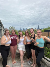 Six friends in activewear holding drinks on a Chicago rooftop patio with the city skyline and cloudy sky in the background.