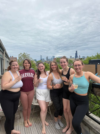 Six women in athletic wear standing on a Chicago rooftop balcony, smiling and holding drinks with the city skyline and overcast sky in the background.