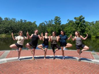 Six people in activewear doing tree pose together on a brick lakeside promenade — outdoor yoga/group fitness on a sunny day with lush green trees reflected in the calm water.