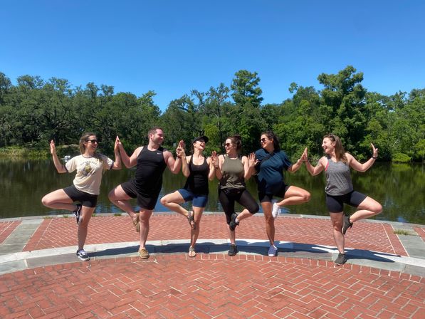 Six people in activewear doing tree pose together on a brick lakeside promenade — outdoor yoga/group fitness on a sunny day with lush green trees reflected in the calm water.