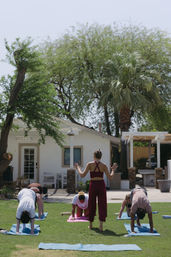 Sunny outdoor group yoga class on a green lawn in a residential backyard, instructor guiding students on mats in downward dog and kneeling poses in front of a white house with a palm tree.