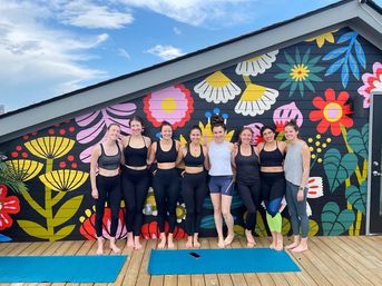 Eight women in workout clothes standing barefoot on a wooden rooftop deck with yoga mats, smiling in front of a vibrant floral mural under a blue sky — outdoor group yoga/fitness session