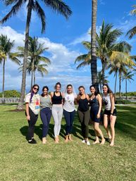 Seven friends, including a bride-to-be in a veil and sash, posing barefoot on a sunny coastal lawn with tall palm trees and a bright blue sky