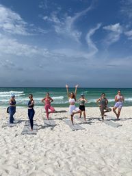 Seven people practicing balancing yoga poses on mats on a white-sand beach with turquoise ocean waves and a bright blue sky with wispy clouds.