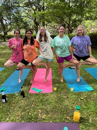 Five women in colorful tees and shorts practicing tree pose on yoga mats in a shady city park, with yoga mats, drinks and a bottle on the grass—outdoor group yoga session vibe.