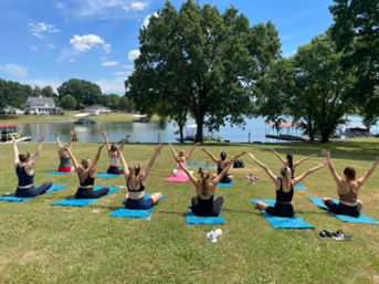 Lively outdoor yoga class on blue mats at a sunny lakeside park, participants seated with arms raised toward the water and trees.