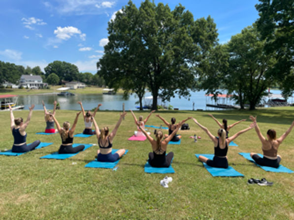 Lively outdoor yoga class on blue mats at a sunny lakeside park, participants seated with arms raised toward the water and trees.