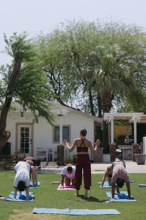 Outdoor group yoga class on a sunny suburban backyard lawn, instructor in maroon guiding students on blue and pink mats beneath palm and shade trees in front of a white house.