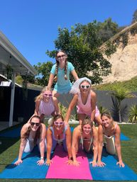 Playful group of seven women in pastel activewear and sunglasses forming a three-level human pyramid on colorful yoga mats in a sunny backyard with a coastal bluff and palm plants; one woman wearing a bridal veil.