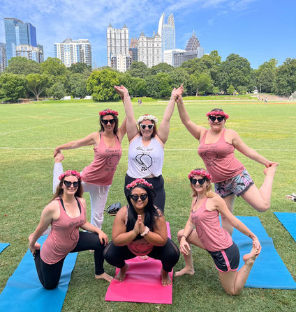 Outdoor group yoga: six people in flower crowns on colorful mats striking poses on a sunny green lawn in an urban park with trees and a city skyline of skyscrapers in the background.