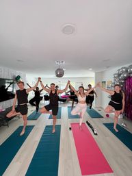 Indoor group yoga class in a bright home studio — adults holding hands in tree pose on teal and pink mats beneath a disco ball, with silver balloons and party decor.