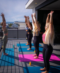 Five women in activewear practicing rooftop yoga on colorful mats, arms stretched overhead toward a blue sky with an urban skyline beyond the railing and glasses of orange juice beside their mats.