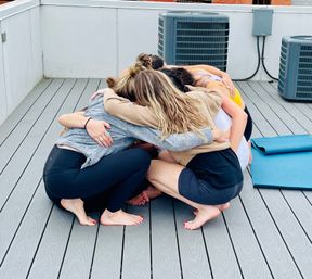 Five people in activewear huddled barefoot in a cozy rooftop yoga circle on a gray composite deck next to rolled blue yoga mats and rooftop HVAC units.