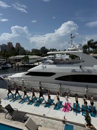 Sunlit outdoor yoga class on mats along a palm-lined marina dock beside a large white luxury yacht, waterfront homes and dramatic cumulus clouds overhead.