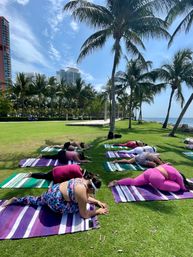 Outdoor yoga class on colorful striped mats on a sunny seaside lawn lined with palm trees, people in stretching poses with ocean and high-rise skyline in the background.