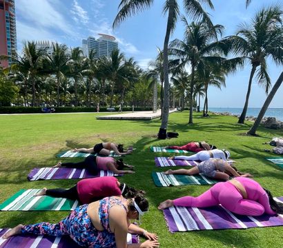 Outdoor yoga class on colorful striped mats on a sunny seaside lawn lined with palm trees, people in stretching poses with ocean and high-rise skyline in the background.