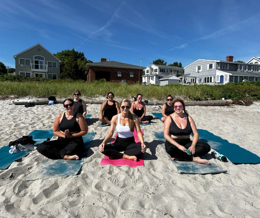 Sunlit group beach yoga: seven women seated on colorful mats on a sandy shore with seaside houses and blue sky in the background.