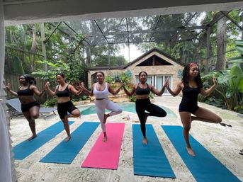 Group yoga: five women in athletic wear hold hands and balance in tree pose on colorful mats by a backyard pool under a screened lanai, with tropical plants and a stone cabana in the background.