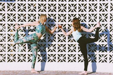 Two women in standing bow yoga pose clinking drinks outdoors against a decorative white breeze-block wall, sunny moment