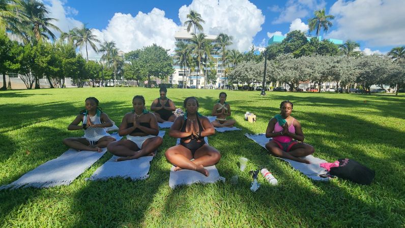 Group of women practicing outdoor yoga meditation on blankets in a sunny tropical urban park with palm trees, green lawn, and blue sky