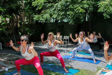 Group of women in colorful leggings laughing and doing a playful wide-legged yoga squat on mats during an outdoor backyard garden yoga/fitness class
