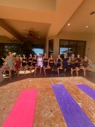Sunlit lineup of women in athletic wear holding tree pose on a covered patio behind pink and purple yoga mats, staged for an outdoor yoga/fitness retreat