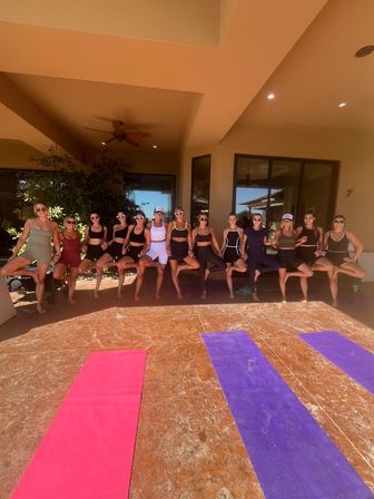 Sunlit lineup of women in athletic wear holding tree pose on a covered patio behind pink and purple yoga mats, staged for an outdoor yoga/fitness retreat