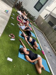 Outdoor group yoga session in a suburban backyard: women lying on colorful mats on artificial turf doing relaxed stretches, water bottles and a wooden fence in the background.