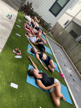 Outdoor group yoga session in a suburban backyard: women lying on colorful mats on artificial turf doing relaxed stretches, water bottles and a wooden fence in the background.