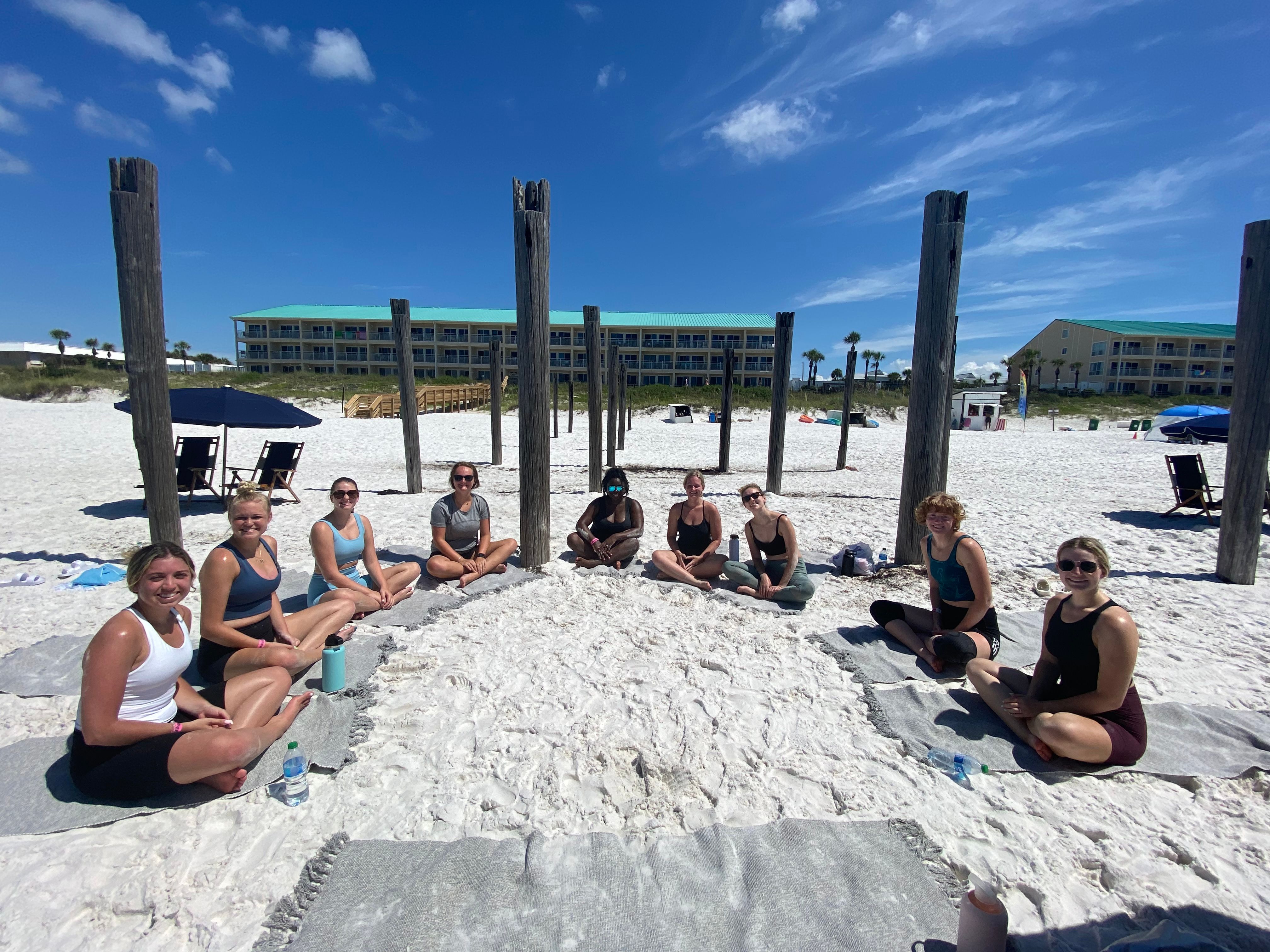 Group of people seated in a meditation/yoga circle on a sunny white-sand beach framed by weathered wooden pilings, bright blue sky and beachfront condo buildings in the background