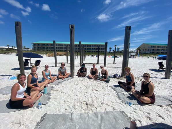 Group of people seated in a meditation/yoga circle on a sunny white-sand beach framed by weathered wooden pilings, bright blue sky and beachfront condo buildings in the background